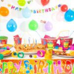 Colorful birthday party table setup with a pie with lit candles, fruit slices, drinks, party hats, and decorations.